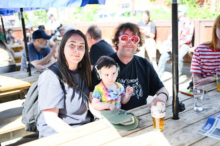 Three people sit together at a wooden picnic table in a sunny outdoor patio at Dandy during Sled Island 2026. On the left, a woman with long dark hair and a striped shirt smiles at the camera. In the center, a small child wearing colorful tie‑dye clothes and protective headphones sits on her lap, holding a green cap. On the right, a man with curly hair and large pink heart‑shaped sunglasses grins, wearing a black T‑shirt with white text. Glasses of beer and water rest on the table, and other people are seated in the background under blue umbrellas, creating a relaxed, cheerful atmosphere.