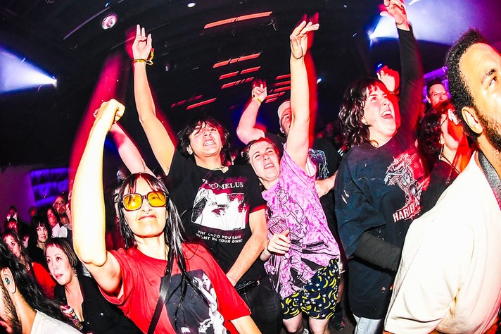During Sled Island 2026, a vibrant crowd at a concert cheers with arms raised under bright, colorful stage lights. People in the front row smile and shout, wearing graphic T‑shirts and casual clothes. One person in a red shirt and yellow sunglasses stands out, raising a fist in excitement. The lighting casts vivid red, purple, and blue tones across the scene, capturing the high‑energy atmosphere of live music and collective joy.