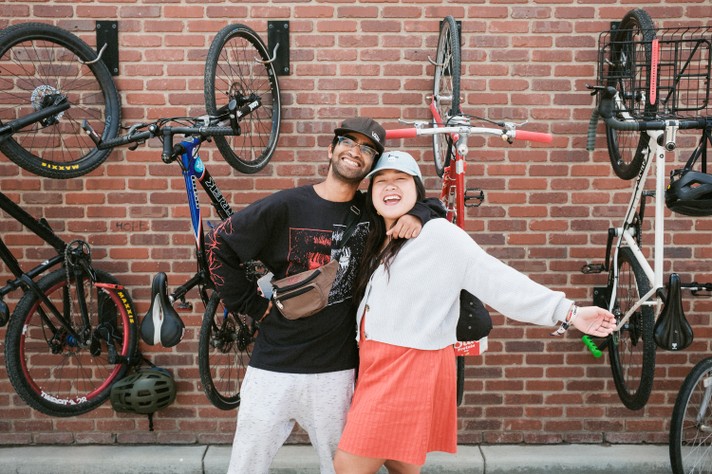During Sled Island 2026, two friends stand smiling in front of a brick wall with several bicycles mounted vertically. The person on the left wears a black long-sleeve shirt with a red graphic, light pants, a brown crossbody bag, and a dark cap, while the person on the right wears a light sweater, orange skirt, and pale blue cap. They pose cheerfully with arms around each other, radiating joy and camaraderie. The background of hanging bikes and helmets adds an urban, active vibe to the scene.