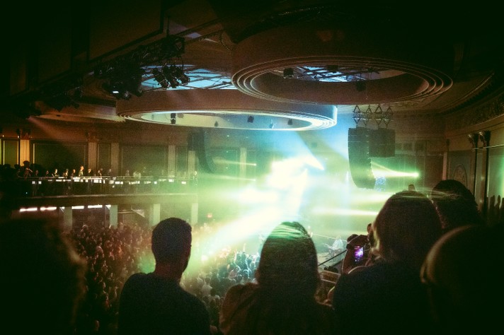 At The Palace Theatre during Sled Island 2026, the packed venue glows with beams of green, yellow, and blue light cutting through haze as a crowd watches the performance below. The photo is taken from an upper balcony, showing silhouettes of audience members in the foreground and rows of people filling both levels of the venue. The circular ceiling design and suspended speakers frame the stage, where bright lights create a dramatic, immersive atmosphere that captures the energy of live music.