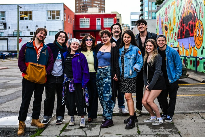 During Sled Island 2026, a group of ten young adults stands close together in an urban alleyway, smiling at the camera. They wear colourful, casual outfits—denim jackets, patterned pants, and sneakers—showing a mix of personal styles. Behind them, a bright mural with cartoonish characters and vivid colours decorates the wall, adding a lively, artistic backdrop to the scene. The group’s relaxed poses and cheerful expressions convey friendship and creative energy.